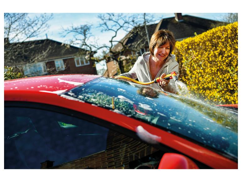 Frau wäscht rotes Auto mit Gartenschlauch und Spritzpistole, Haus und Hecke im Hintergrund.