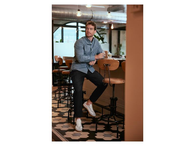 Ein Mann in einem hellblauen Hemd, schwarzen Hosen und weißen Sneakern sitzt an einer Bar.