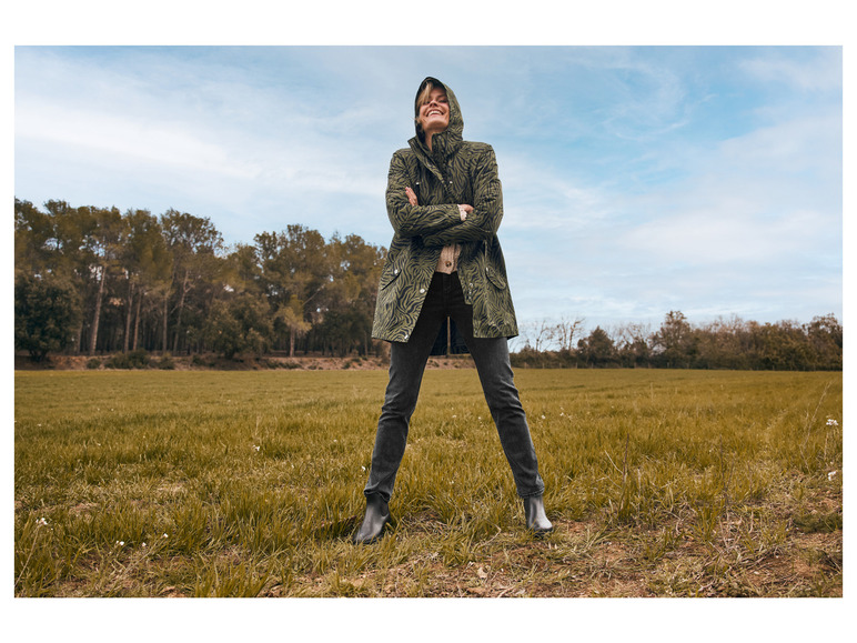 Eine Frau trägt einen grünen Regenmantel mit Tigermuster und schwarze Stiefel auf einem Feld.