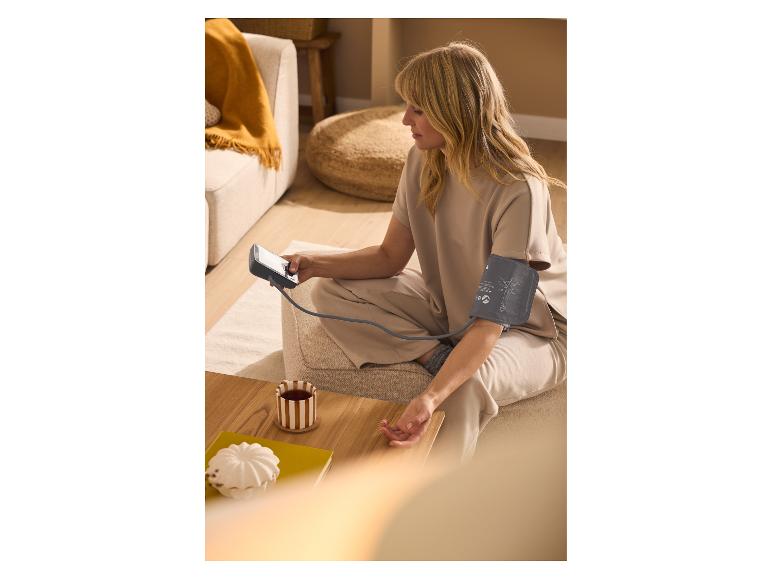 Frau misst Blutdruck mit Oberarm-Blutdruckmessgerät, sitzt auf dem Boden mit einer Tasse Tee.