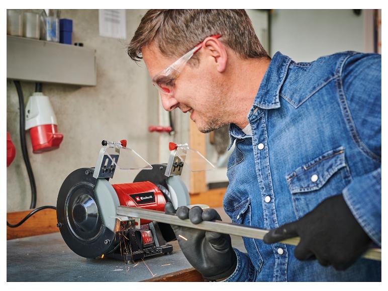 Mann mit Schutzbrille und Handschuhen schleift Metall an einem Tischschleifer.