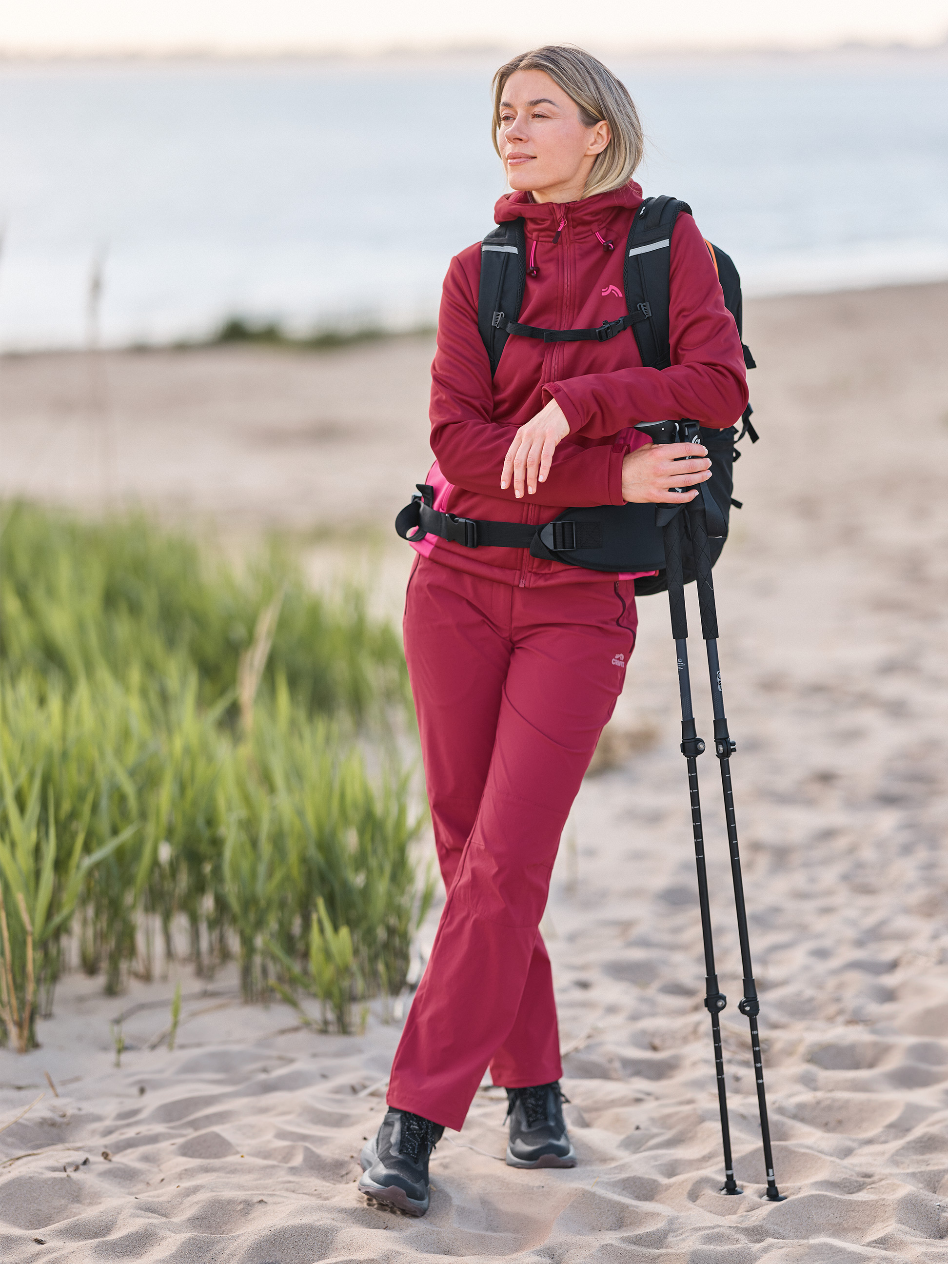 Frau in Wanderkleidung und Rucksack am Strand, hält Trekkingstöcke.