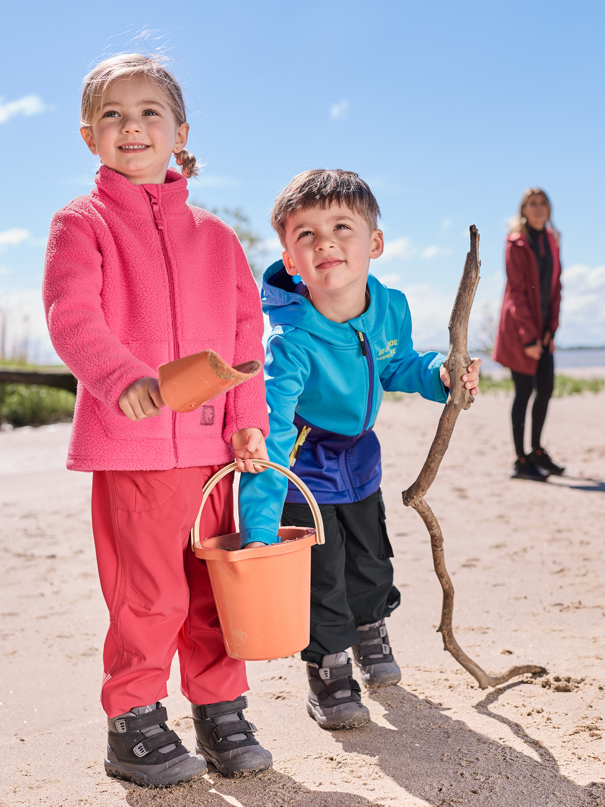 Zwei Kinder spielen am Strand, tragen rosa und blaue Fleecejacken und wasserdichte Hosen.