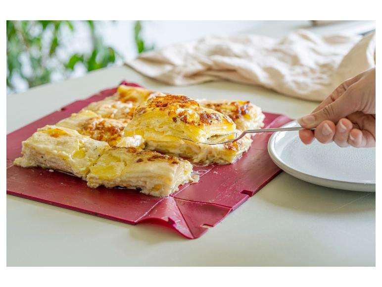 Eine Hand serviert eine Portion Kartoffelgratin von einem roten Tablett auf einen Teller.