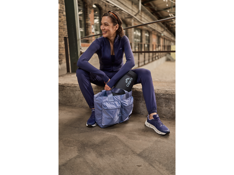 Eine Frau in lila Sportkleidung sitzt mit einer Sporttasche und einer Gymnastikmatte da.