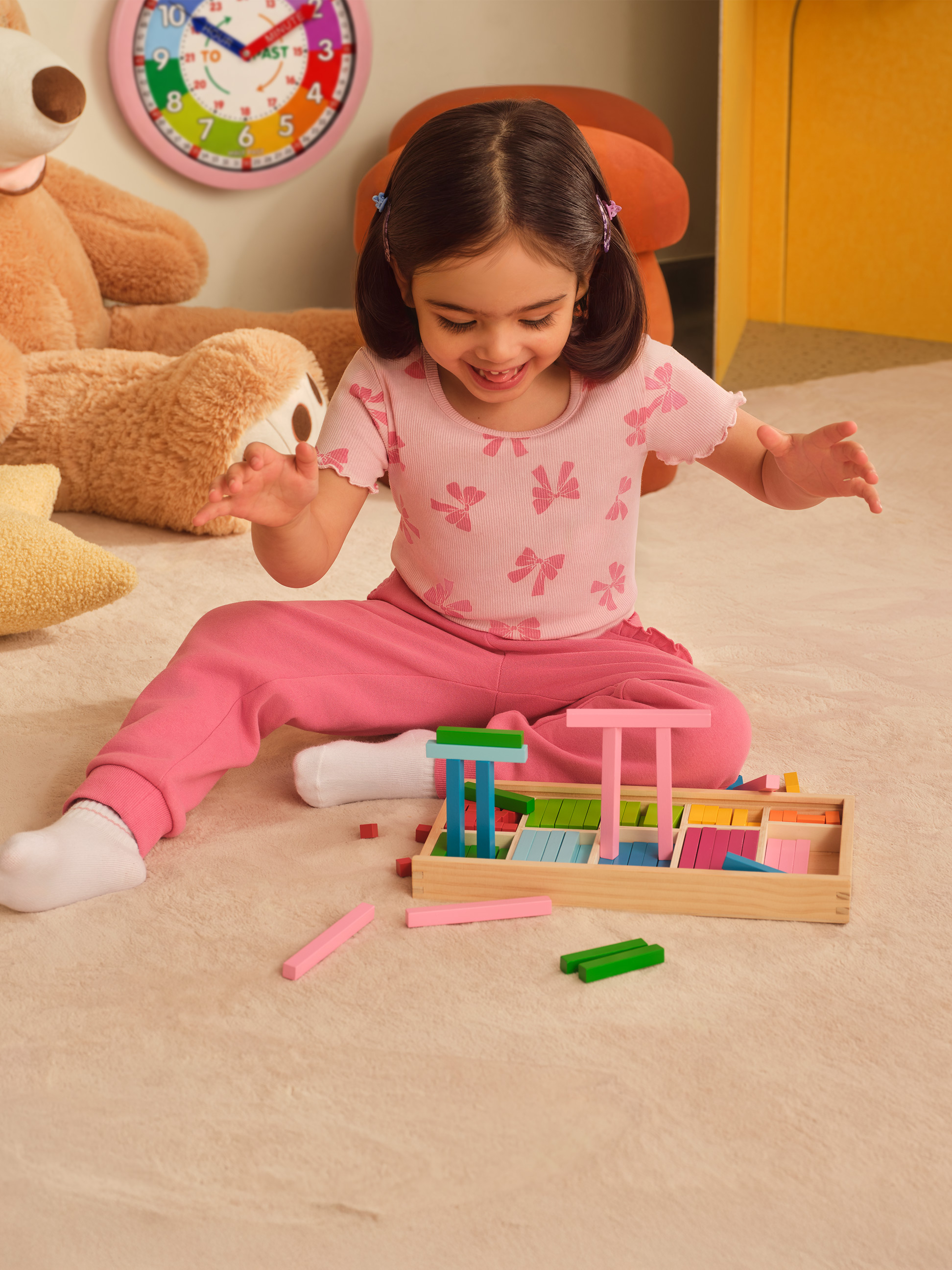Lächelndes Mädchen spielt mit bunten Holzklötzen auf einem Teppich, mit Teddybär und Uhr im Hintergrund.