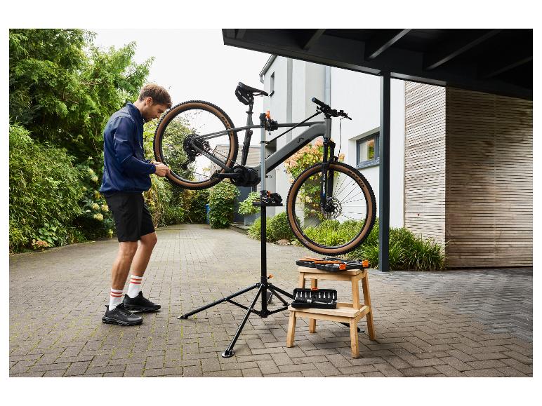 Mann in blauer Jacke und schwarzen Shorts repariert ein Fahrrad auf einem Ständer mit Werkzeugen auf einem Hocker.