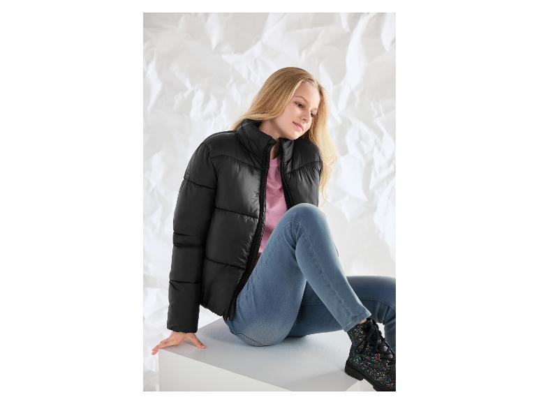 Junges Mädchen in schwarzer Pufferjacke, rosa T-Shirt und Jeans sitzt auf einer weißen Box.