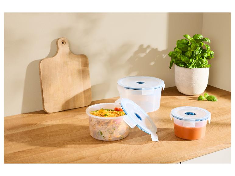 Drei transparente Lebensmittelbehälter mit Essen und ein Schneidebrett auf einer Küchenarbeitsplatte.