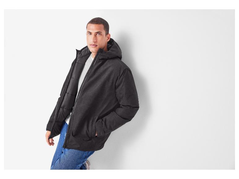Mann in schwarzer Daunenjacke und Jeans lehnt an einer weißen Wand.
