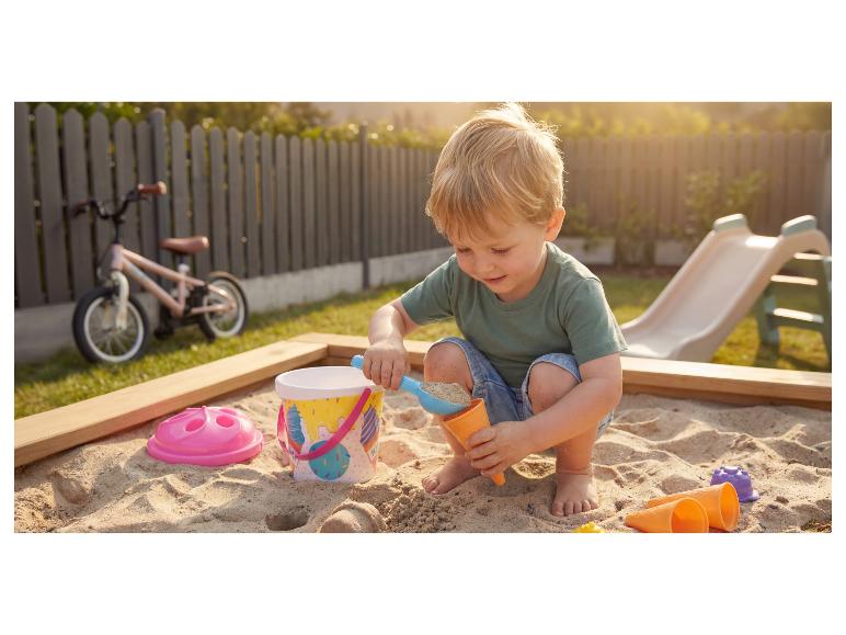 Kleinkind spielt im Sandkasten mit Eimer und Schaufel, mit Fahrrad und Rutsche im Hintergrund.