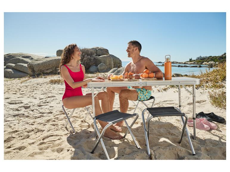 Paar genießt ein Strandpicknick mit Klapptisch und -stühlen, Obst und einer Wasserflasche.