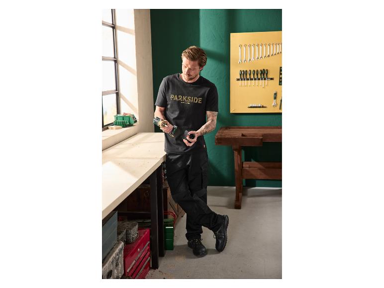 Mann in schwarzem T-Shirt und Hose hält Bohrmaschine in Werkstatt.