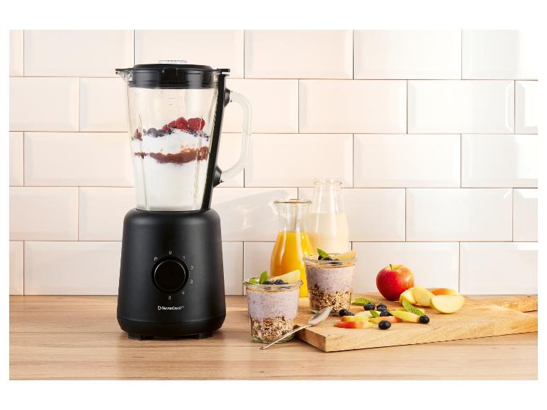 Schwarzer Mixer mit Beeren und Joghurt, daneben Gläser mit Frühstück und Obst.