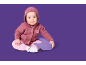 Baby in rosa Kapuzenstrickjacke und rosa Hose, sitzt auf einem lila Kissen.