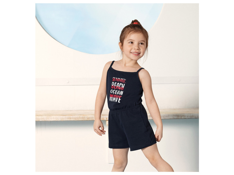 Ein Mädchen trägt einen blauen Overall mit dem Schriftzug 'Summer Beach Ocean Wave'.