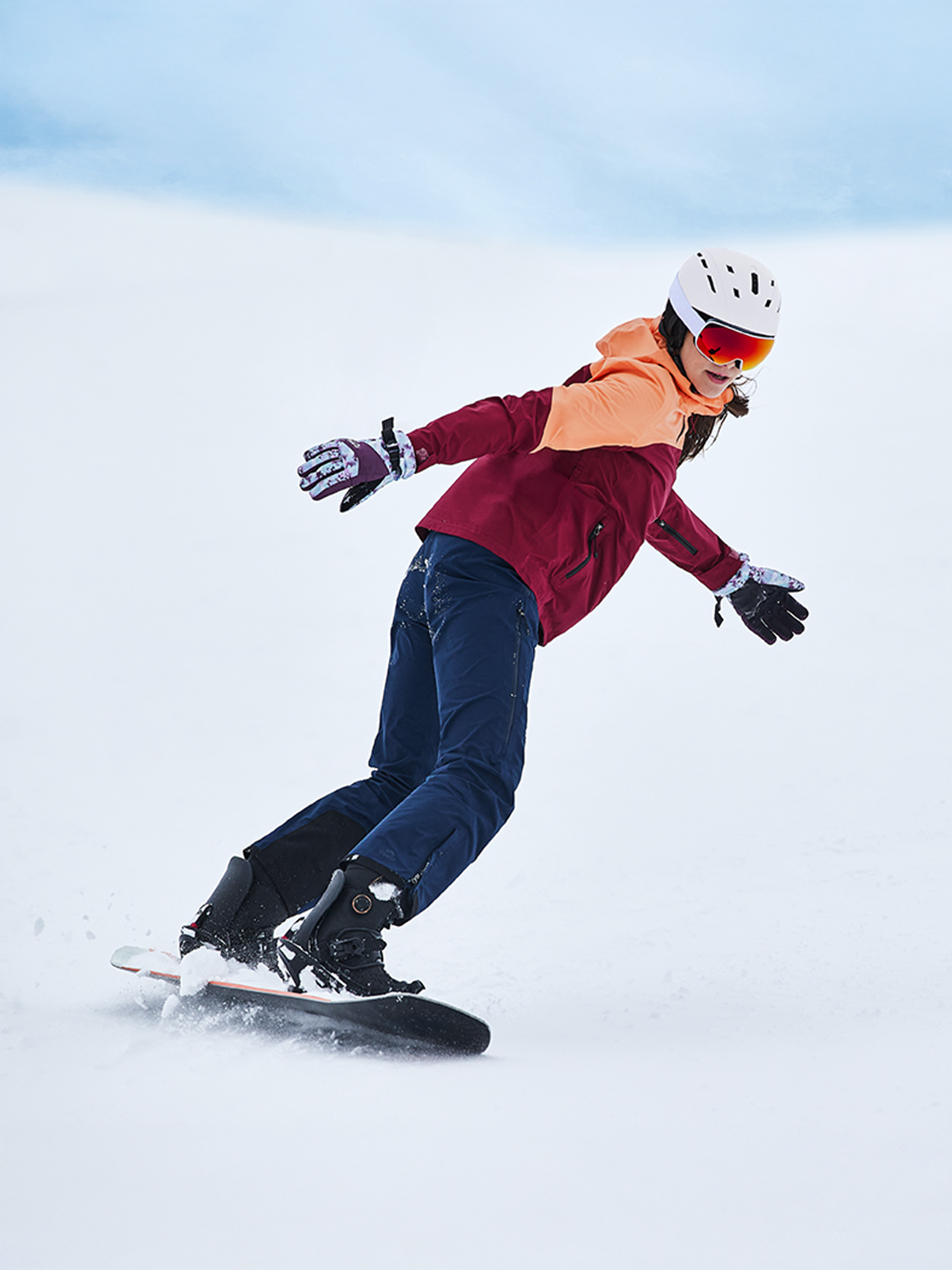 Ein Snowboarder in einer rot-orangen Jacke, blauen Hosen und einem weißen Helm im Schnee.
