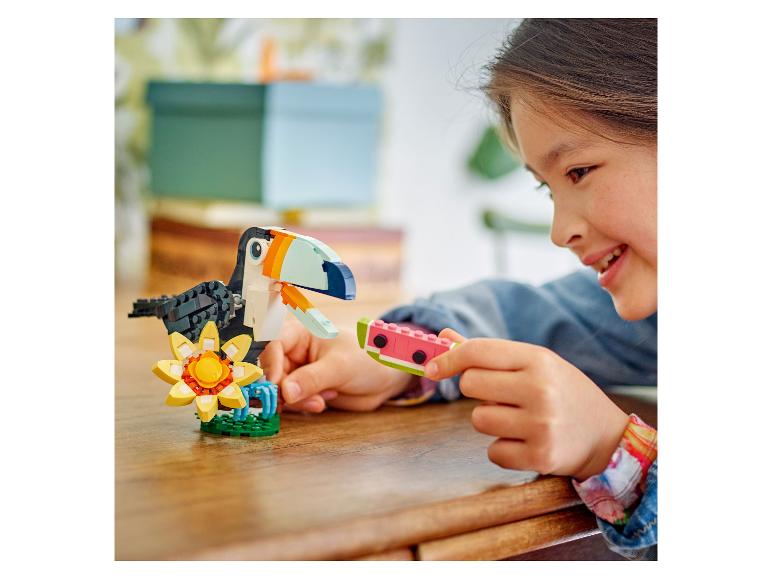 Ein Mädchen lächelt, während es ein LEGO Wassermelonenstück in der Nähe eines LEGO Tukans und einer Blume hält.