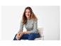 Frau in grauem Zopfstrickpullover und Jeans sitzt lächelnd auf einem Stuhl.