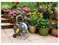 Grauer und gelber Gartenschlauchwagen auf einer sonnigen Terrasse mit bunten Topfpflanzen.