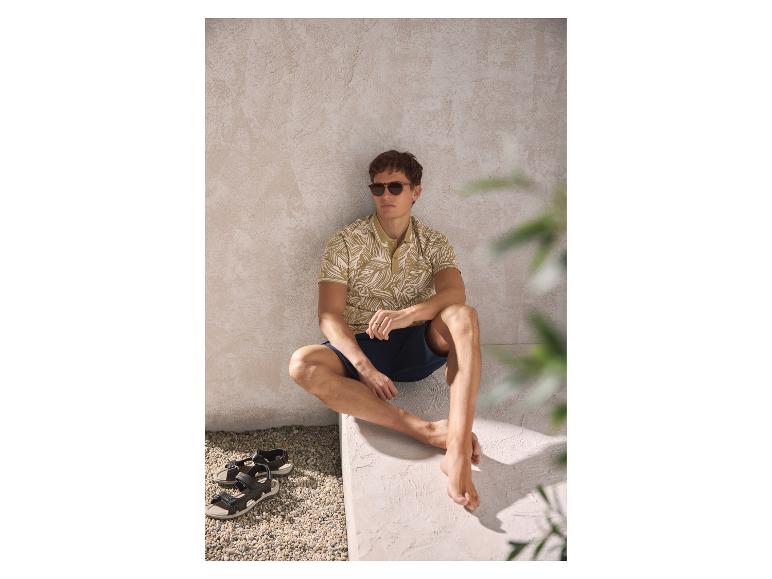 Mann in gemustertem Poloshirt und Shorts, sitzt mit Sandalen daneben.