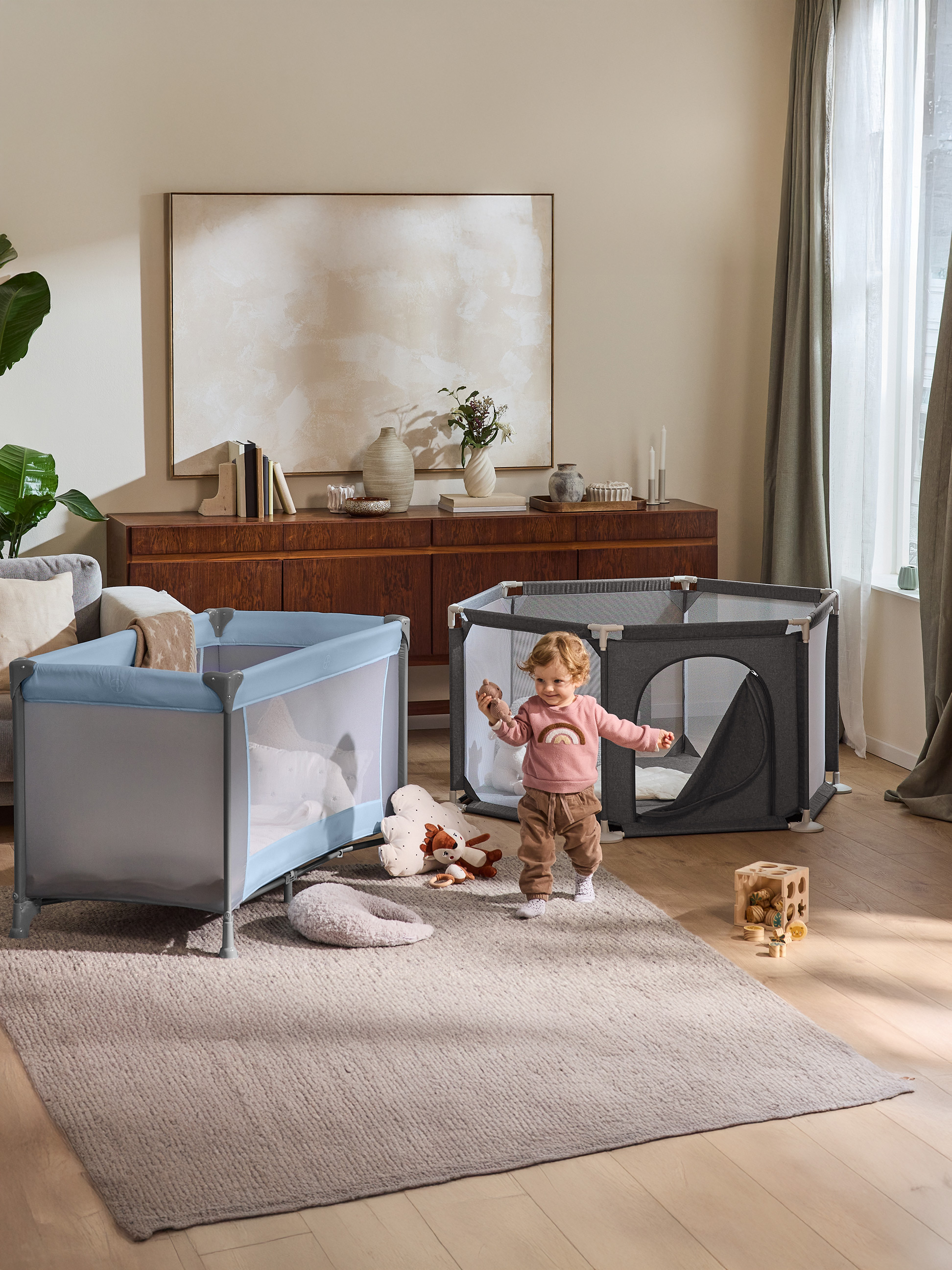 Kleinkind spielt im Wohnzimmer mit Laufstall und Reisebett.