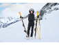 Eine Frau in schwarzer Skikleidung posiert mit orangefarbenen Skiern auf einem verschneiten Berghang.