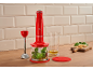 Roter Stabmixer mit Basilikum, Öl und Käse auf einer Holztheke.