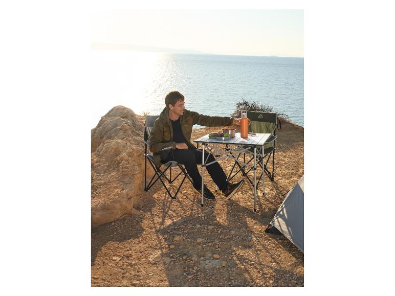 Mann sitzt an Campingtisch mit Stühlen, neben Zelt, mit Meerblick.