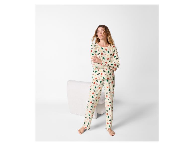 Frau in cremefarbenem Pyjama mit Weihnachtsmuster, barfuß stehend neben einem weißen Stuhl.