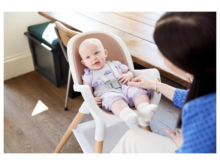 Lächelndes Baby in lila Kleidung sitzt im Hochstuhl, ein Erwachsener hält seine Beine.