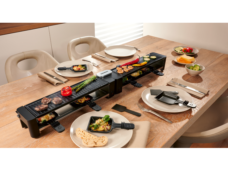 Langer Raclette-Grill auf einem gedeckten Holztisch mit Essen und Tellern