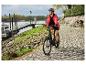 Frau fährt E-Bike am Flussufer, trägt rote Jacke und Helm.