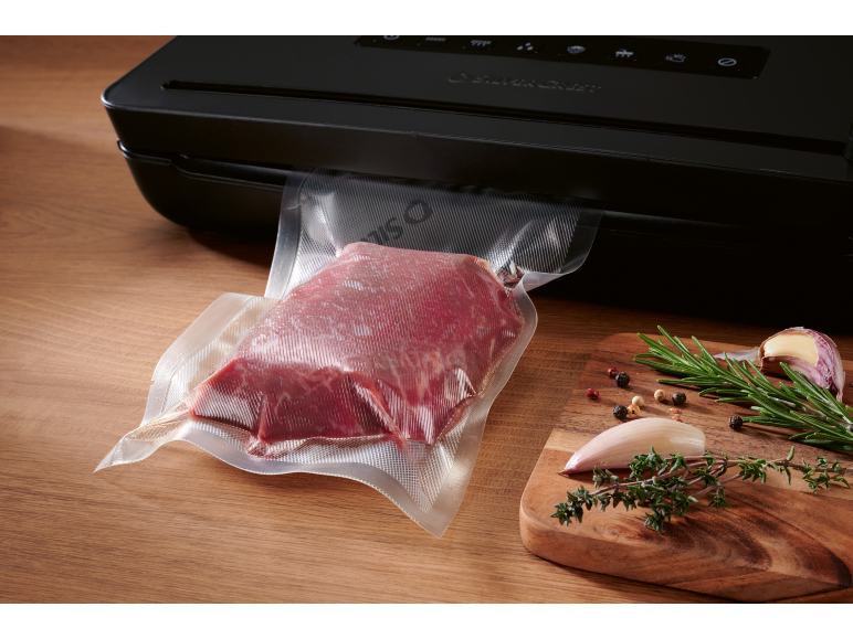 Vakuumverpacktes Fleisch mit Gewürzen auf einem Holzbrett neben einem Vakuumiergerät.