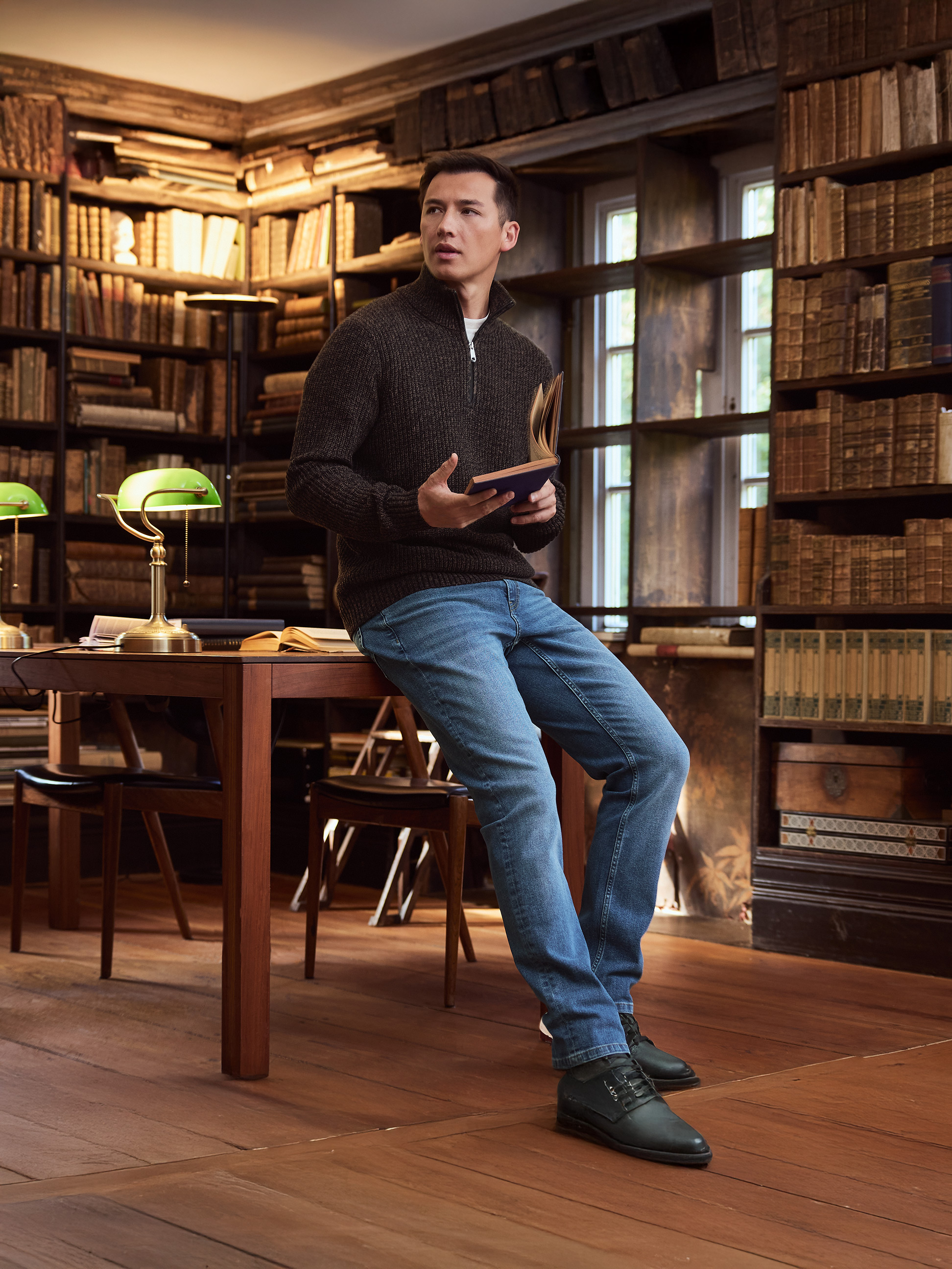 Mann in braunem Pullover und Jeans liest ein Buch in einer Bibliothek.