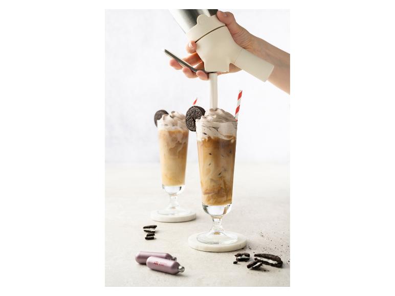 Hand sprüht Schlagsahne auf Eiskaffee mit Keksen und Gaskartuschen auf dem Tisch.