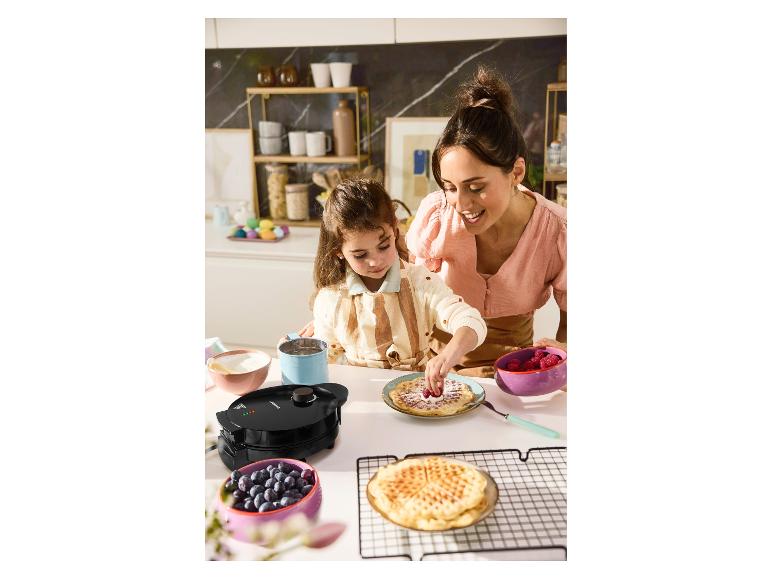 Mutter und Tochter dekorieren Waffeln mit Beeren und Blaubeeren, neben einem Waffeleisen.