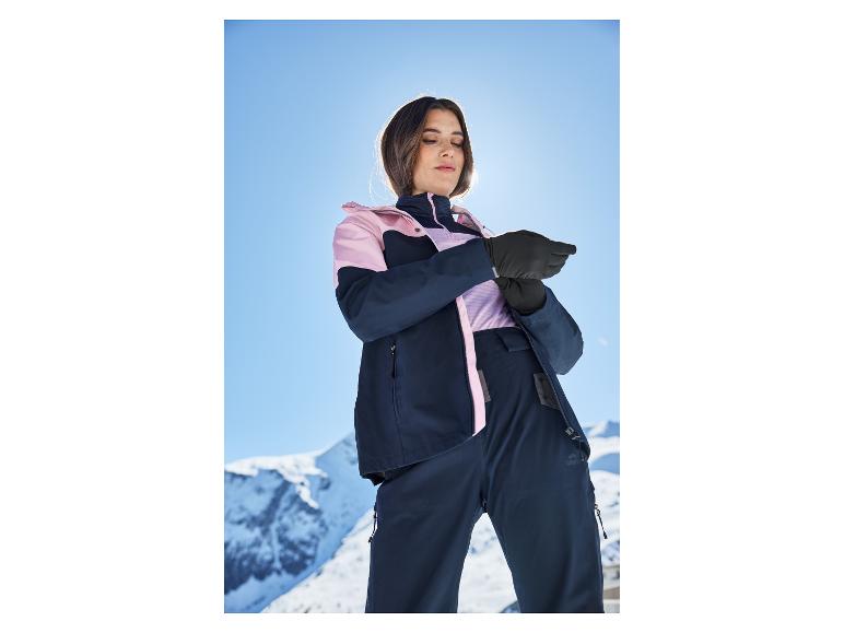 Frau in Skijacke und -hose, die ihre Handschuhe auf einer sonnigen Piste anpasst.