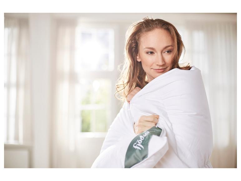 Eine Frau, eingehüllt in eine weiße Fjödur-Bettdecke in einem hellen Raum.