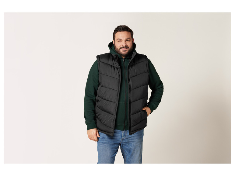 Mann trägt eine schwarze Steppweste, einen grünen Hoodie und blaue Jeans.