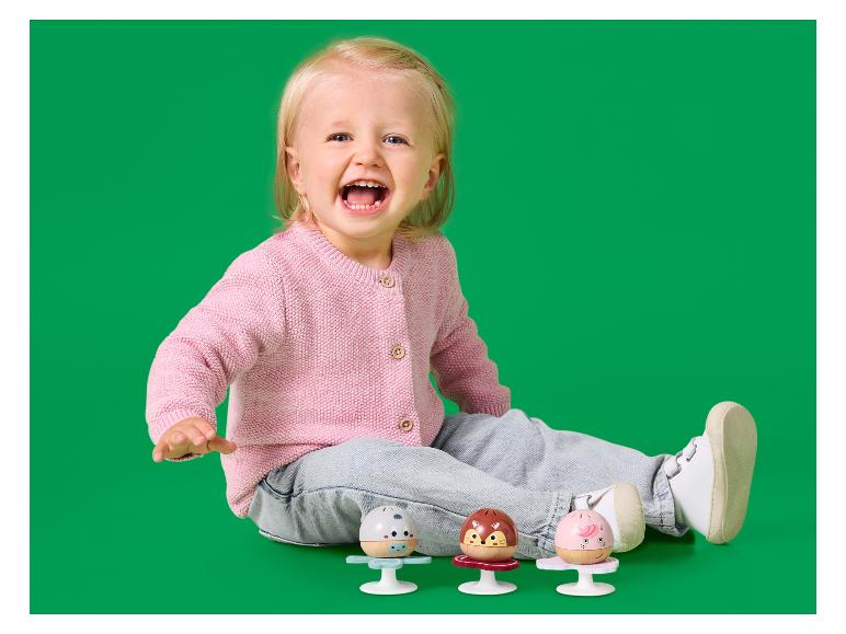 Glückliches Baby in rosa Strickjacke und Jeans, spielt mit drei bunten Rasseln.