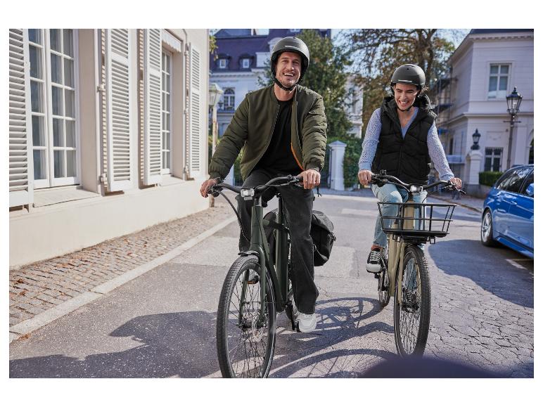 Ein Paar fährt Fahrrad auf einer Stadtstraße mit Gebäuden und einem blauen Auto im Hintergrund.