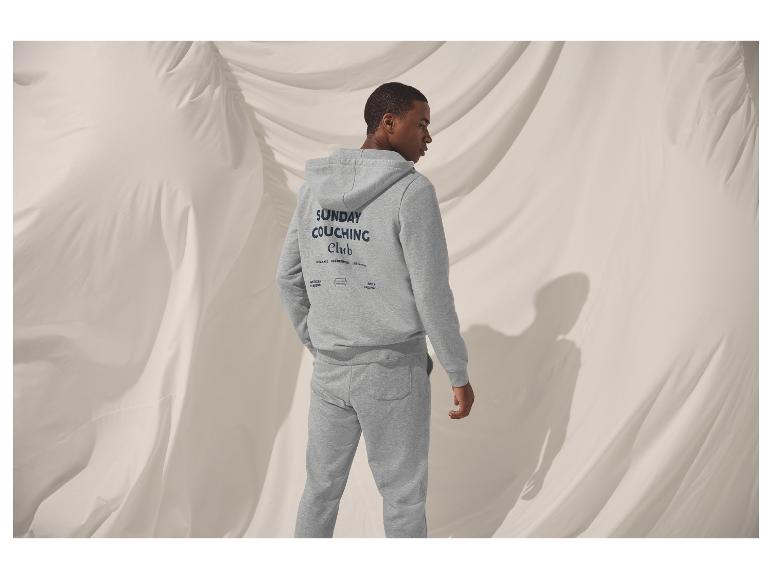 Mann in grauem Kapuzenpullover und Jogginghose mit dem Text „SUNDAY COUCHING Club“ auf dem Rücken.