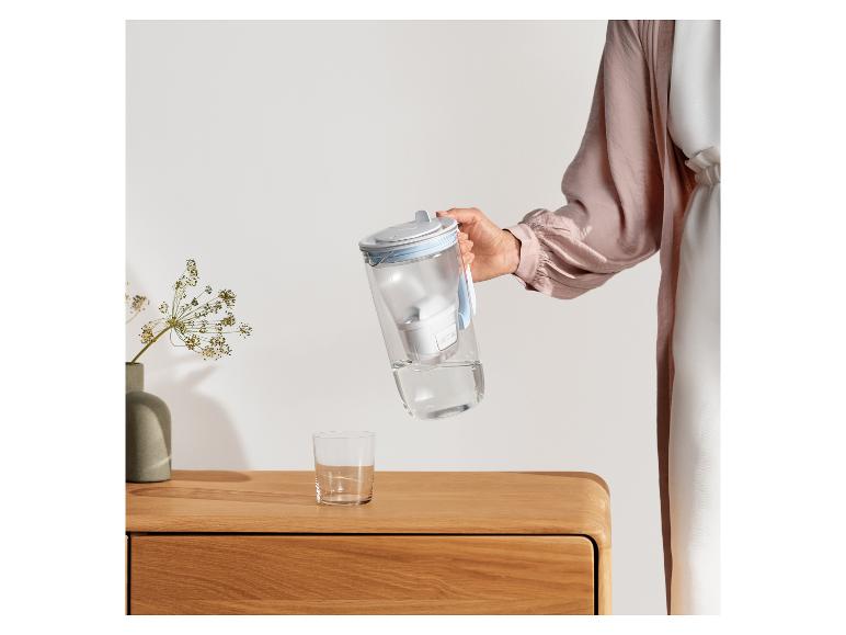 Eine Person gießt Wasser aus einem Filterkrug in ein Glas auf einem Holznachttisch.