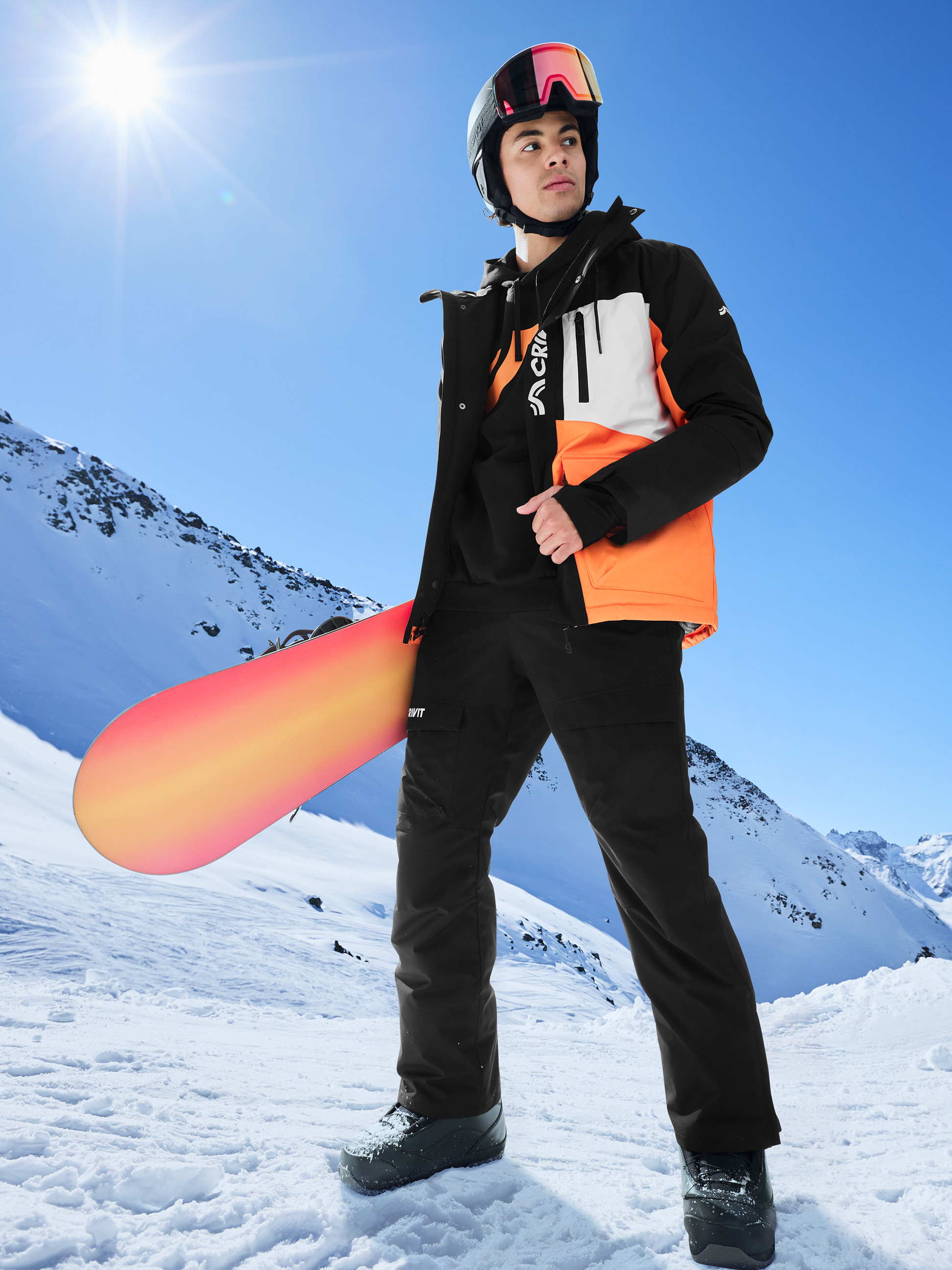 Mann in schwarz-orangefarbener Skijacke, schwarzer Skihose und Helm mit Snowboard im Schneegebirge.