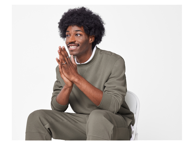 Mann mit Afro sitzt auf einem Stuhl in einem olivgrünen Sweatshirt und Jogginghose.