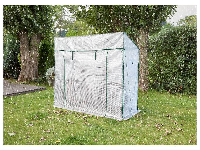 Ein transparentes Gewächshaus in einem Garten mit Bäumen und Gras, bei fallendem Regen.