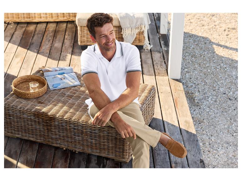 Mann in einem weißen Poloshirt und beigen Hosen sitzt auf einem Rattanstuhl.