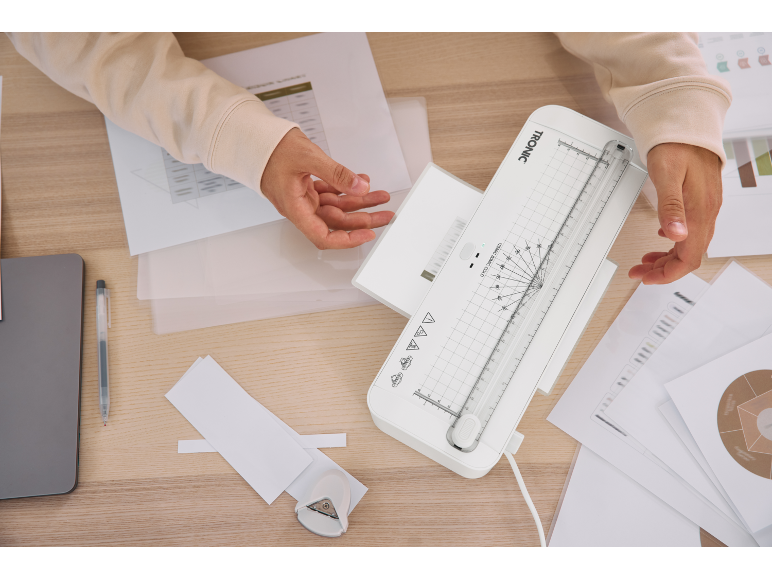 Eine Person laminiert und schneidet Papier mit einem weißen Laminiergerät auf einem Holztisch.