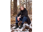 Frau in Winterstiefeln und Jacke sitzt auf einem Baumstumpf im verschneiten Wald.